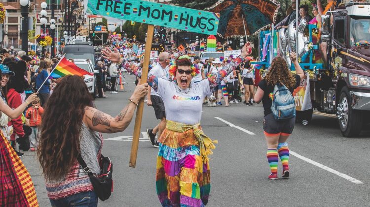 Lynelle in a rainbow striped skirt wearing a Camosun College shirt with a rainbow logo joyfully running up to woman holding "Free Mom Hugs" sign.